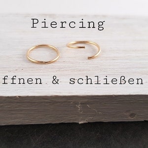 May include: Two gold-colored hoop earrings, one open and one closed, on a white wooden surface. The text "Piercing &ouml;ffnen & schlie&szlig;en" is written below the earrings.
