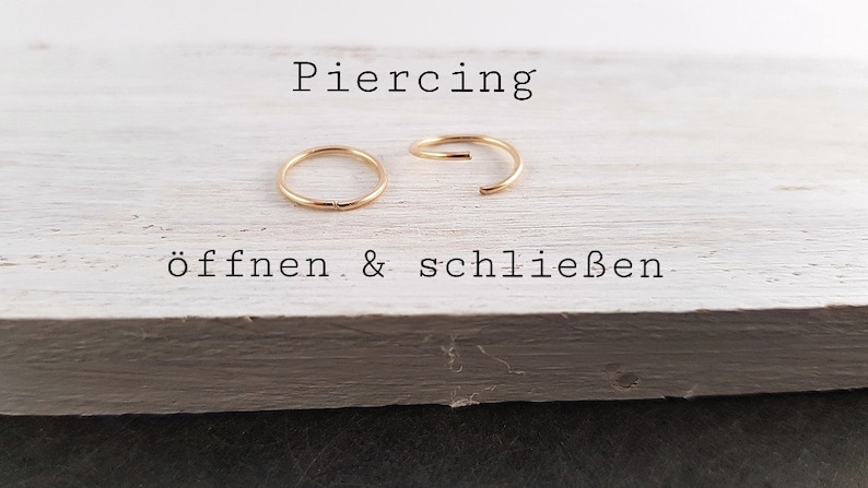 May include: Two gold-colored hoop earrings, one open and one closed, on a white wooden surface. The text "Piercing &ouml;ffnen & schlie&szlig;en" is written below the earrings.