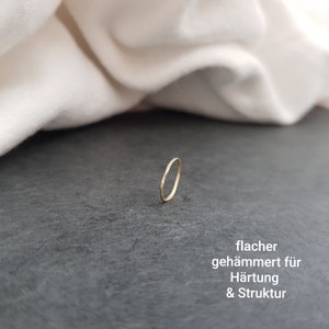 May include: A thin, hammered gold ring. The ring is flat and has a smooth, polished finish. "flacher geh&auml;mmert f&uuml;r H&auml;rtung & Struktur" is written in German below the ring.