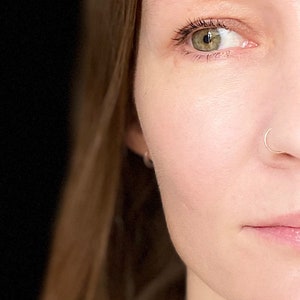 May include: Close-up of a person's face with green eyes and a nose ring. The person has light skin and is looking to the side.