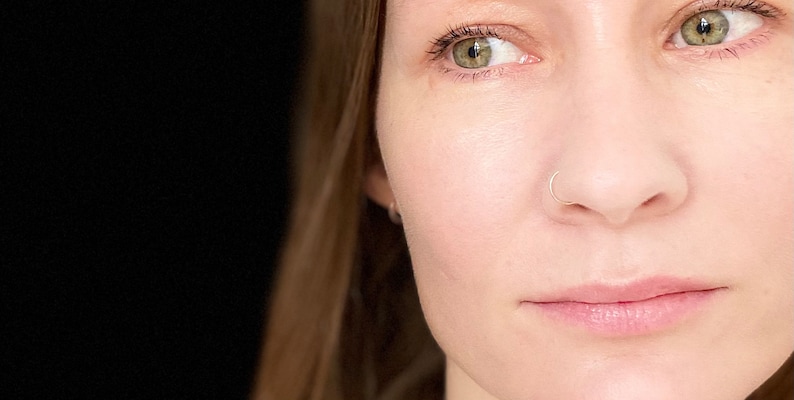 May include: Close-up of a person's face with green eyes and a nose ring. The person has light skin and is looking to the side.