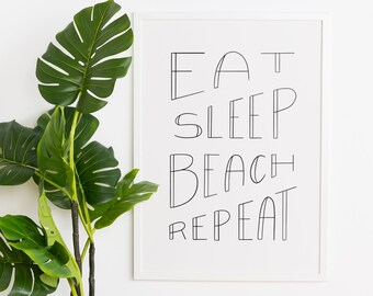 May include: A white framed print with black text that reads "Eat Sleep Beach Repeat" hanging on a white wall with a green potted plant in front of it.