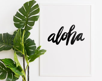 May include: A white framed print with the word "aloha" written in black cursive script. A potted green plant with large leaves sits on a wooden surface in front of the print.