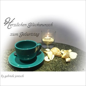 Könnte beinhalten: Eine tealfarbene Teetasse und Untertasse mit einer brennenden Kerze in einem Glasgefäß und Muscheln auf einer dunkelgrauen Oberfläche. Der Text "Herzlichen Glückwunsch zum Geburtstag" ist in schwarzer Kursivschrift über der Kerze geschrieben.