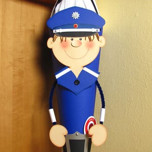 Könnte beinhalten: Eine kegelförmige Schultüte im Design eines Polizisten, mit blauer Uniform, passender Polizeimütze mit silbernem Abzeichen, lächelndem Gesicht, dunkelblauen Pfeifenputzer-Armen und einem rot-weißen runden Schild mit der Aufschrift HALT.