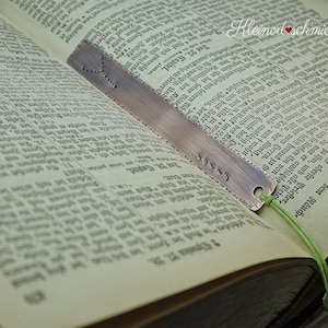 Könnte beinhalten: Ein Kupfer-Lesezeichen mit einem dekorativen Rand und einem grünen Band. Das Lesezeichen liegt offen auf einem Buch mit dem Text "Kleinod schmiede" sichtbar.