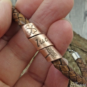 Może przedstawiać: Bransoletka ze skóry brązowej z miedzianą zawieszką z napisem "szczęście" i motywem koniczyny czteroliściennej oraz ważki.