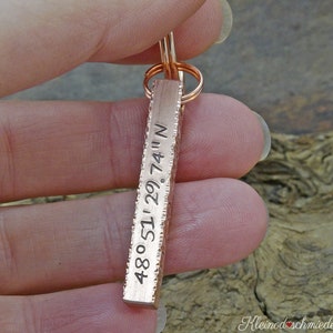 May include: A copper-coloured rectangular keychain pendant with engraved coordinates: 48°51'29.74"N. The pendant has a textured edge and is attached to a copper-coloured split ring.