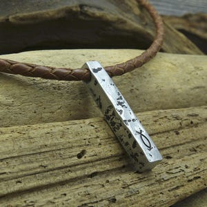 Könnte beinhalten: Eine silberne Anhänger-Halskette mit einem geflochtenen braunen Lederband. Der Anhänger ist eine rechteckige Stange mit einer abgenutzten Oberfläche und einem eingravierten Fischsymbol.