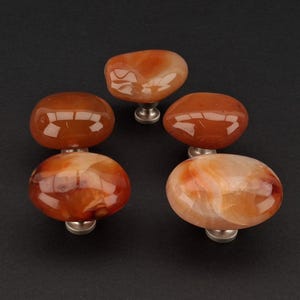 Könnte beinhalten: Fünf polierte, ovale Schrankknöpfe. Die Knöpfe bestehen aus orangefarbenem und braunem Achatstein und sind auf silberfarbenen Metallsockeln montiert. Die Knöpfe sind vor einem dunklen Hintergrund angeordnet und zeigen ihre natürlichen Muster und Farben.