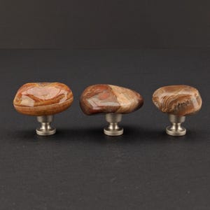 Könnte beinhalten: Drei dekorative Schrankknöpfe mit Oberflächen aus Naturstein in Braun-, Rot- und Beigetönen. Jeder Knopf ist auf einer gebürsteten Nickelbasis montiert. Die Knöpfe haben eine glatte, polierte Oberfläche und variieren leicht in Form und Muster.