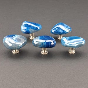 Könnte beinhalten: Fünf dekorative Schrankknöpfe mit einem polierten blau-weißen Achatstein-Design. Jeder Knopf ist auf einer gebürsteten silbernen Basis montiert. Die Knöpfe haben unterschiedliche Formen, darunter runde und ovale Formen.