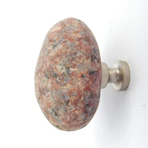 Könnte beinhalten: Einzigartiger Schrankknauf mit Naturstein-Design. Der Knauf hat eine abgerundete, längliche Form mit einer Mischung aus roten, grauen und weißen Farben. Er ist an einer gebürsteten Metallbasis befestigt und bietet eine rustikale, aber moderne Ästhetik.