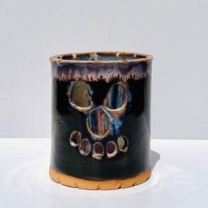 May include: A black ceramic Halloween-themed candle holder with a skull face design. The candle holder has a brown base and a dark blue and brown rim. A Halloween garland with orange pumpkins, black bats, and white ghosts hangs in the background.