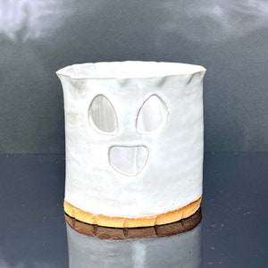 May include: White ceramic ghost-shaped candle holder with cut-out eyes and a smiling mouth. The holder has a light brown rim.