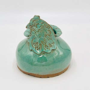 May include: A teal ceramic vase with a decorative floral design on the lid. The vase has a rounded body and a slightly flared opening.