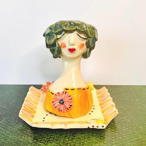 May include: A ceramic planter shaped like a woman's head with green hair, red lips, and rosy cheeks. The planter is wearing a yellow dress with a pink flower and sits on a matching tray with a scalloped edge.