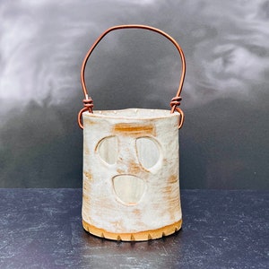 May include: White ceramic candle holder with a ghost face design and a copper wire handle. The holder is sitting on a dark gray surface.