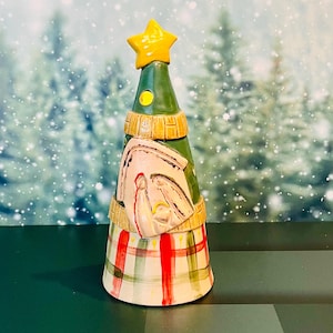 May include: A ceramic Christmas tree figurine with a yellow star on top. The tree is green with a cream-colored band and a plaid base in red, green, and white. The tree has a nativity scene on the front.