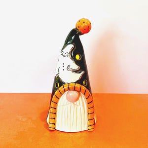 May include: A Halloween gnome figurine with a black hat featuring a ghost design and a pumpkin on top. The gnome has a white beard and an orange arched frame. Decorative cutouts of ghosts, a pumpkin, and a bat hang in the background.