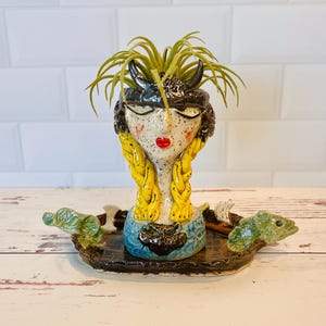 May include: A whimsical ceramic planter shaped like a woman's face, featuring yellow braids and a black horned helmet. The planter holds green plants and sits on a tray with green turtle-like figures. The face has red lips and a blue dress.