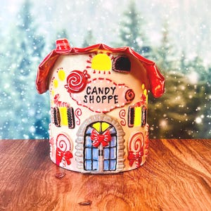 May include: A ceramic candy shoppe house figurine with a red roof, featuring painted details of lollipops, cookies, and a central door with a red bow. The front facade displays the text CANDY SHOPPE in black lettering on a light pink background.
