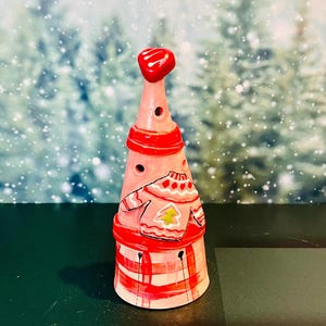 May include: A pink ceramic Christmas tree-shaped candle holder with a red heart on top. The tree has a red and white striped base and a sweater design with a yellow Christmas tree. The background is a snowy forest.