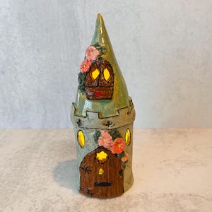 May include: A whimsical, hand-painted ceramic castle figurine. The tower features a green conical roof, a brown door, and windows that appear to glow. Pink flowers and green foliage adorn the exterior, adding a touch of fantasy. The castle is a decorative item.