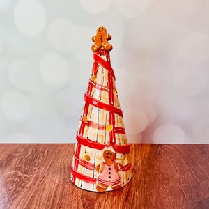 May include: A ceramic Christmas tree with a gingerbread theme. The tree is cone-shaped, decorated with red ribbon, yellow accents, and gingerbread people. The tree is approximately 10 inches tall and sits on a wooden surface.
