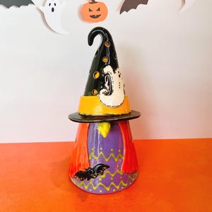 May include: A ceramic Halloween gnome figurine. The gnome wears a black witch hat with a white ghost design, a yellow band, and a black brim. The body is purple with a red cape and a bat. The figurine is on an orange surface.