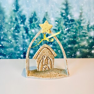 May include: A speckled ceramic nativity ornament featuring a stylized stable structure suspended within an arched frame. A yellow star sits at the top, and a yellow ribbon ties the central piece to the frame. The ceramic has a tan base with brown speckles.