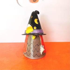 May include: A whimsical Halloween witch figurine. The black witch hat features a candy corn accent and crescent moon details. The witch's body is gray with a zig-zag pattern and a white ghost applique. The figurine has red hair and a purple hat band.
