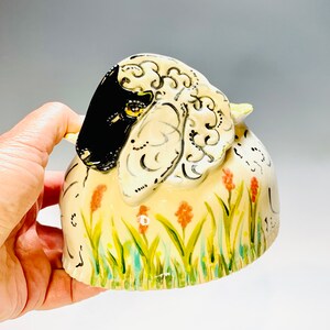 May include: A ceramic sheep figurine with a black face and white wool, standing in a field of green grass with pink flowers. The sheep has a detailed design on its wool.
