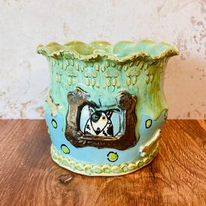 May include: A ceramic planter with a light blue and green glaze. The planter features a brown framed window with a black and white dog illustration. The rim has a scalloped edge. The planter is decorated with yellow circles.