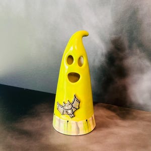 May include: A yellow ceramic ghost-shaped figurine with three facial cutouts and a bat applique. The base features a plaid design in shades of yellow and gray. The figurine is a Halloween decoration.