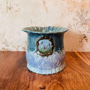 May include: A blue ceramic planter with a scalloped rim and a decorative floral design. The planter features a framed painting of flowers and greenery. The base of the planter has a light blue floral pattern.