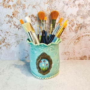 May include: A turquoise ceramic brush holder with a decorative floral design and a gold-framed painting of wildflowers. The holder is filled with various paint brushes of different sizes and shapes, with natural bristles.