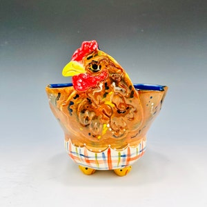 May include: A ceramic chicken-shaped bowl with a brown, yellow, red, and blue painted design. The bowl has three yellow feet and a plaid pattern around the bottom.