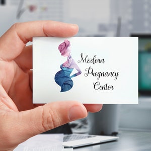 Puede incluir: Una tarjeta de visita sostenida en una mano, con una ilustraci&oacute;n en acuarela de una persona embarazada en tonos azules y morados. La tarjeta dice "Modern Pregnancy Center" en una elegante escritura.