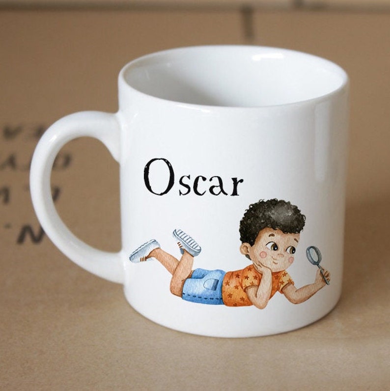 Pode incluir: Caneca de cer&acirc;mica branca com al&ccedil;a preta. A caneca tem uma ilustra&ccedil;&atilde;o de desenho animado de um menino com pele morena e cabelo castanho, vestindo uma camisa azul e laranja, deitado de lado e olhando para uma lupa. A caneca &eacute; personalizada com o nome "Oscar" em texto preto.