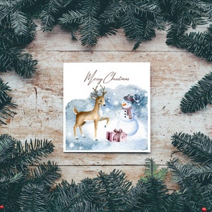 May include: A watercolor Christmas card featuring a deer and a snowman with a red and white striped scarf. The card has a blue background with white snowflakes and the text "Merry Christmas".