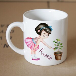 Pode incluir: Caneca de cer&acirc;mica branca com uma ilustra&ccedil;&atilde;o de desenho animado de uma menina com um tutu rosa e cabelo preto, apontando para uma pequena planta em um vaso marrom. O nome da menina, "Rosie", est&aacute; escrito em roxo abaixo da ilustra&ccedil;&atilde;o.