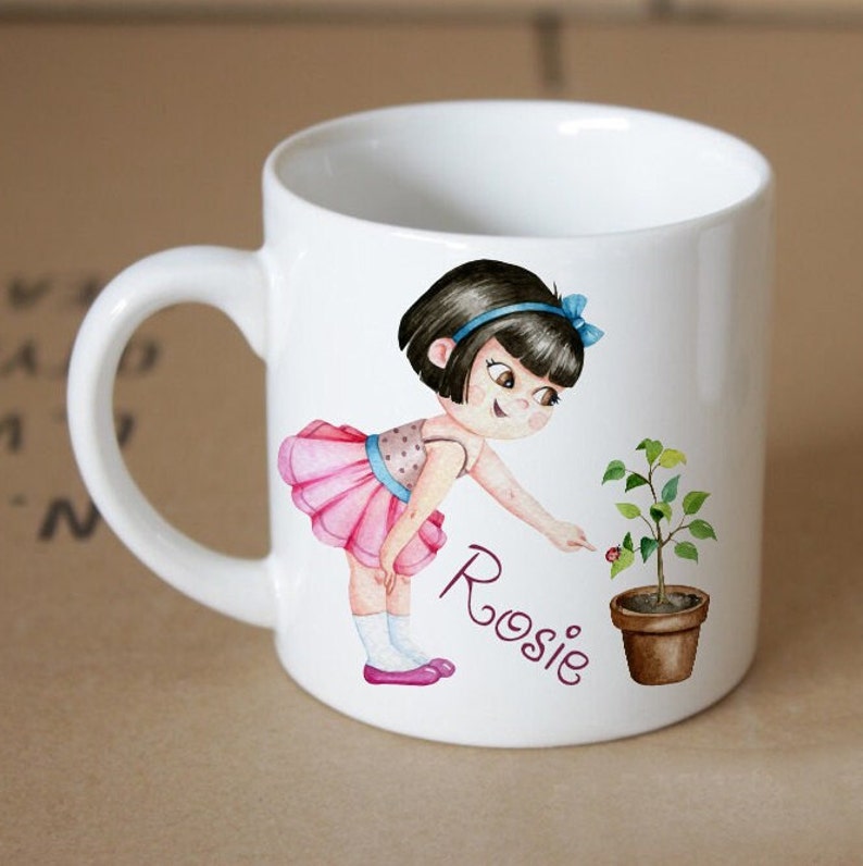 Pode incluir: Caneca de cer&acirc;mica branca com uma ilustra&ccedil;&atilde;o de desenho animado de uma menina com um tutu rosa e cabelo preto, apontando para uma pequena planta em um vaso marrom. O nome da menina, "Rosie", est&aacute; escrito em roxo abaixo da ilustra&ccedil;&atilde;o.