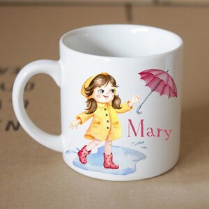Pode incluir: Caneca de cer&acirc;mica branca com uma ilustra&ccedil;&atilde;o em aquarela de uma menina usando um casaco de chuva amarelo e botas de chuva vermelhas, segurando um guarda-chuva vermelho. A menina est&aacute; em p&eacute; em uma po&ccedil;a d'&aacute;gua. A caneca tem o nome "Mary" escrito nela.