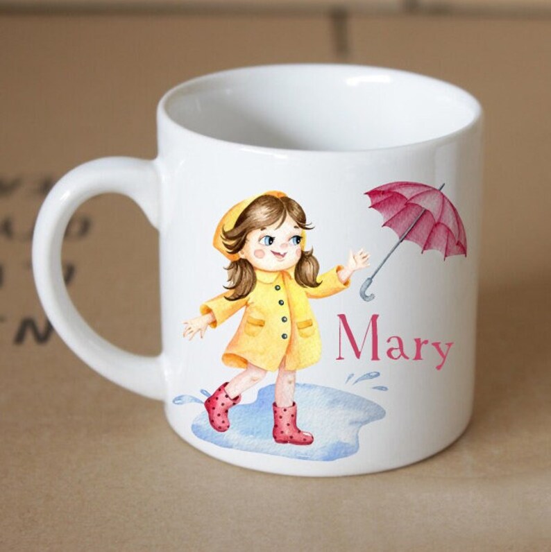 Pode incluir: Caneca de cer&acirc;mica branca com uma ilustra&ccedil;&atilde;o em aquarela de uma menina usando um casaco de chuva amarelo e botas de chuva vermelhas, segurando um guarda-chuva vermelho. A menina est&aacute; em p&eacute; em uma po&ccedil;a d'&aacute;gua. A caneca tem o nome "Mary" escrito nela.