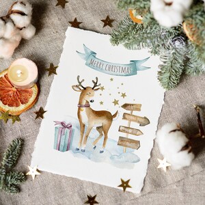 May include: A watercolor illustration of a reindeer standing in the snow with a gift box and wooden signs that read "Cafe", "North Pole", "Toy Shop", and "Santa's Village". The illustration is on a white background with gold stars and the text "Merry Christmas".