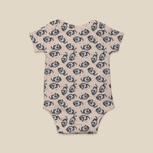 May include: A beige baby bodysuit with a repeating pattern of sleeping sloths. The sloths are depicted in gray and white, creating a playful design. The bodysuit has short sleeves and a simple, practical design.