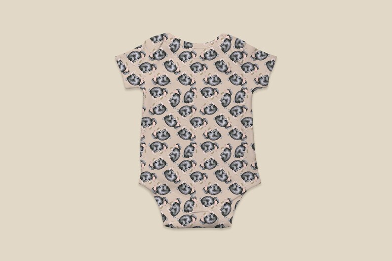May include: A beige baby bodysuit with a repeating pattern of sleeping sloths. The sloths are depicted in gray and white, creating a playful design. The bodysuit has short sleeves and a simple, practical design.