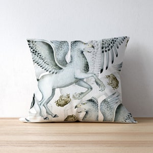 May include: Square throw pillow featuring a watercolor illustration of a griffin, a mythical creature with the body of a lion and the head and wings of an eagle. The design includes shades of gray, white, and brown, with a light background. The pillow is on a wooden surface.