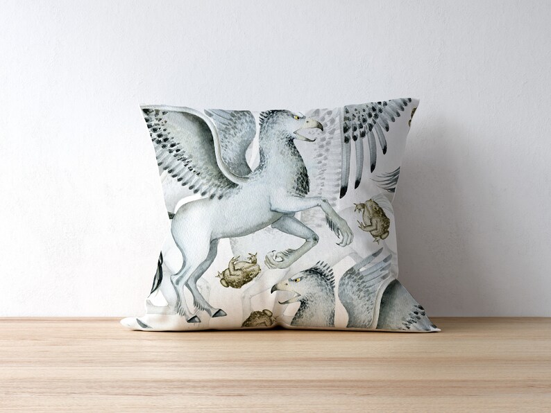 May include: Square throw pillow featuring a watercolor illustration of a griffin, a mythical creature with the body of a lion and the head and wings of an eagle. The design includes shades of gray, white, and brown, with a light background. The pillow is on a wooden surface.
